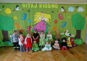 Dzieci z grupy II w wiosennych przebraniach na tle dekoracji.